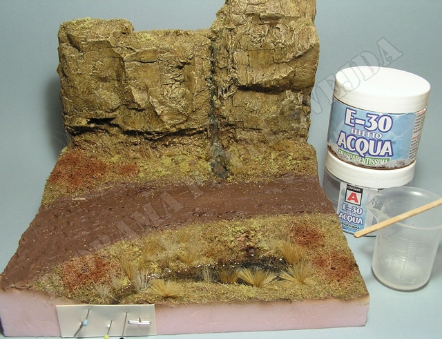 potok.jpg - Koryto potoka nabarven&eacute; tmav&yacute;mi odst&iacute;ny akrylov&yacute;ch barev je pokryto posypem tmav&yacute;ch, skoro čern&yacute;ch odst&iacute;nů jemn&yacute;ch posypov&yacute;ch materi&aacute;lů (Fine turfy od Woodland Scenicsu), zepředu je uzavřen&eacute; plastikovou destičkou a vedle je transparentn&iacute; voda E-30 Aqua firmy Prochima.